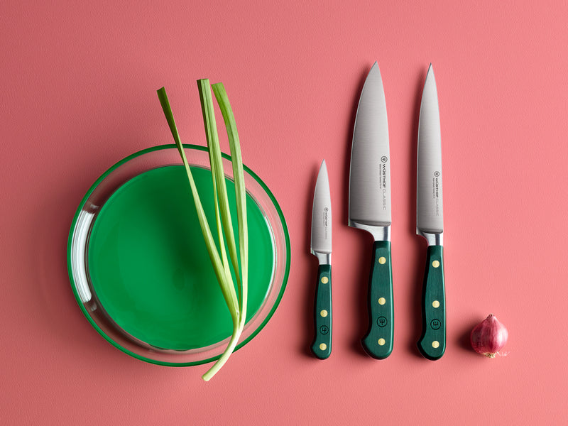 Three knives with green handles on a pink background, next to a green plate with green onions and a small red onion.