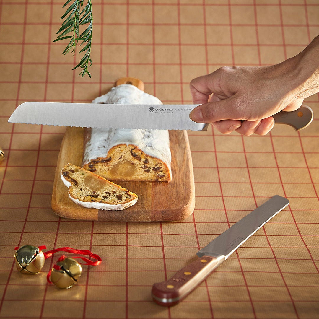 Hand holding a Wusthof bread knife over a loaf of bread on a wooden cutting board with a decorative background.