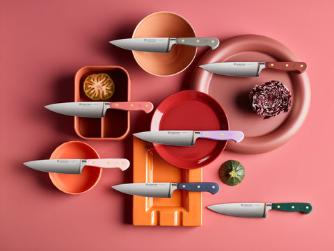 A variety of kitchen knives with colorful handles are arranged on a pink background alongside sliced vegetables and vibrant plates.