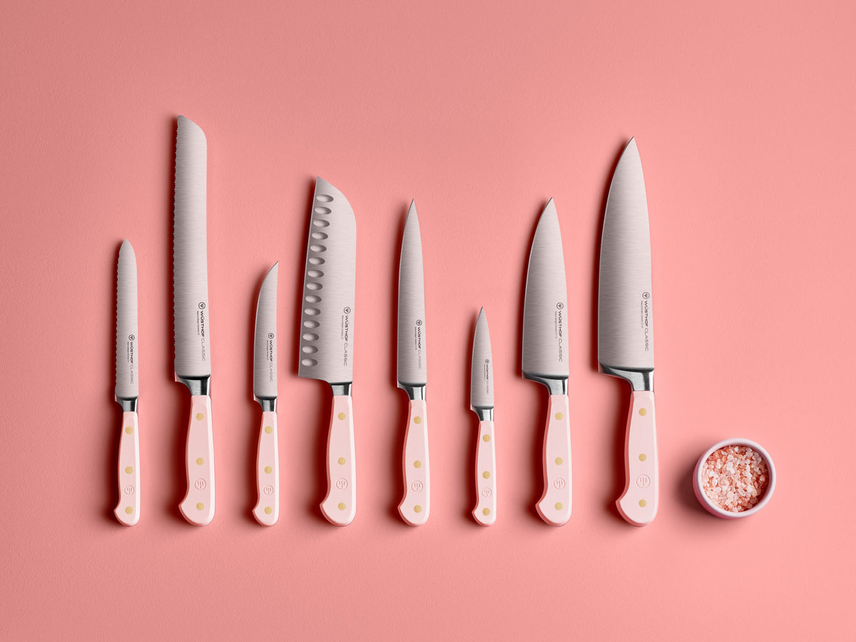Eight kitchen knives with pastel pink handles featuring brass rivets and sharp stainless steel blades, arranged on a soft pink surface alongside a small bowl of pink salt