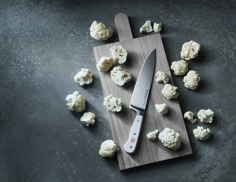 Chef knife with a polished blade and a white handle featuring rivets, placed on a wooden board surrounded by cauliflower florets on a dark surface
