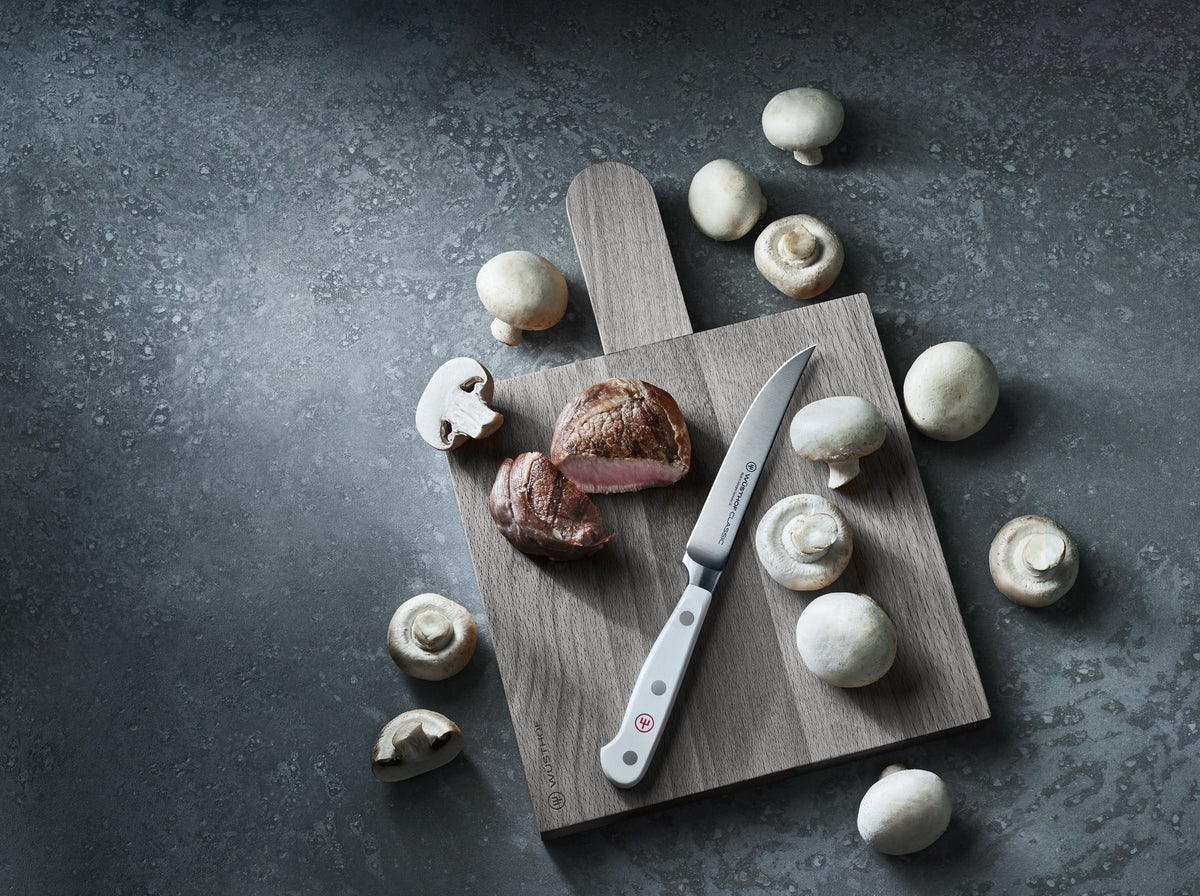 Single steak knife with a white handle resting on a wooden cutting board surrounded by whole and sliced white mushrooms and pieces of cooked meat.