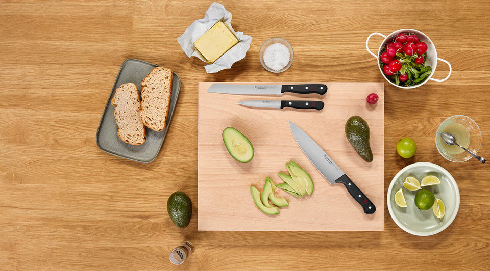 Wooden cutting board with sliced avocados, knives, and a bowl of fruit on a wooden table.