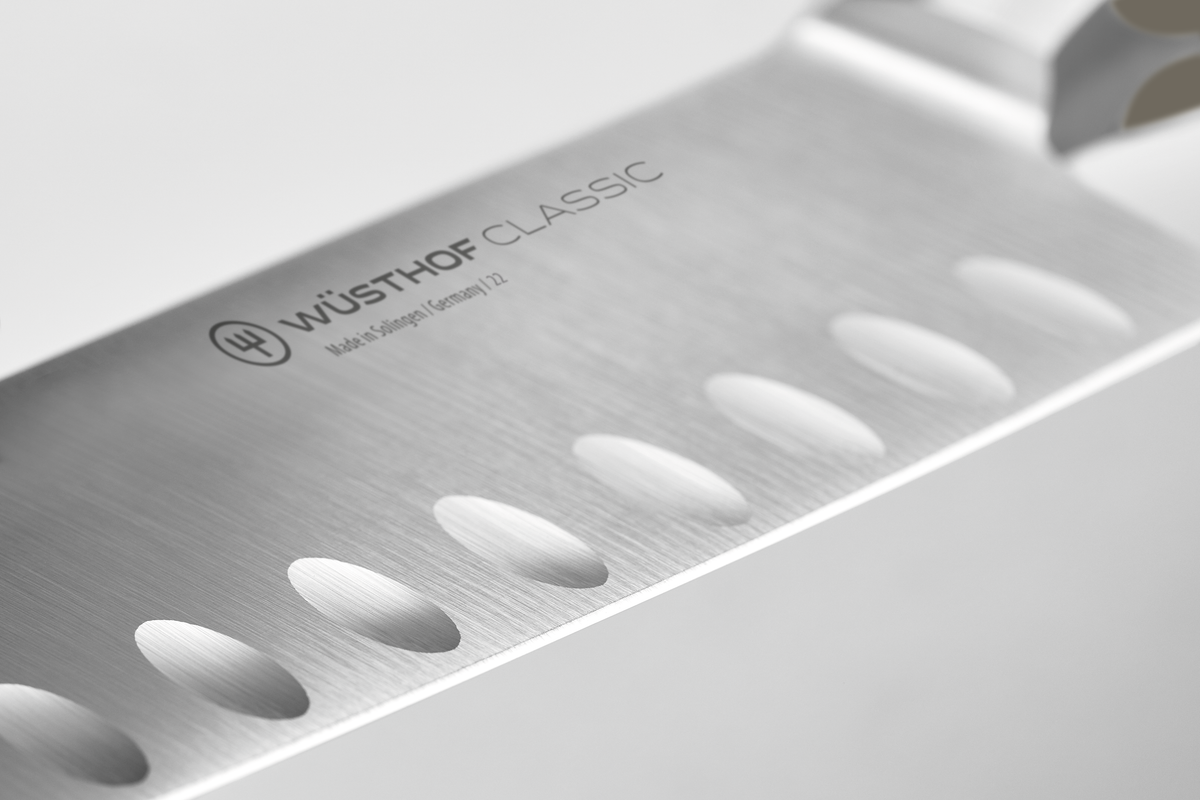 Close-up of a brushed stainless steel kitchen knife blade with hollow-edge indentations and subtle branding on a premium German chef knife.